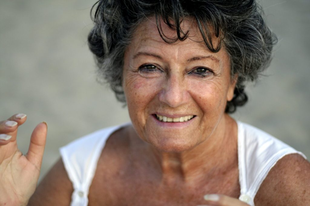 Ritratto di una donna anziana con capelli ricci e grigi, che sorride felicemente mentre alza una mano in segno di saluto.